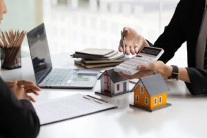 A couple reviews home offer documents at a kitchen table with a stack of paperwork and a laptop.